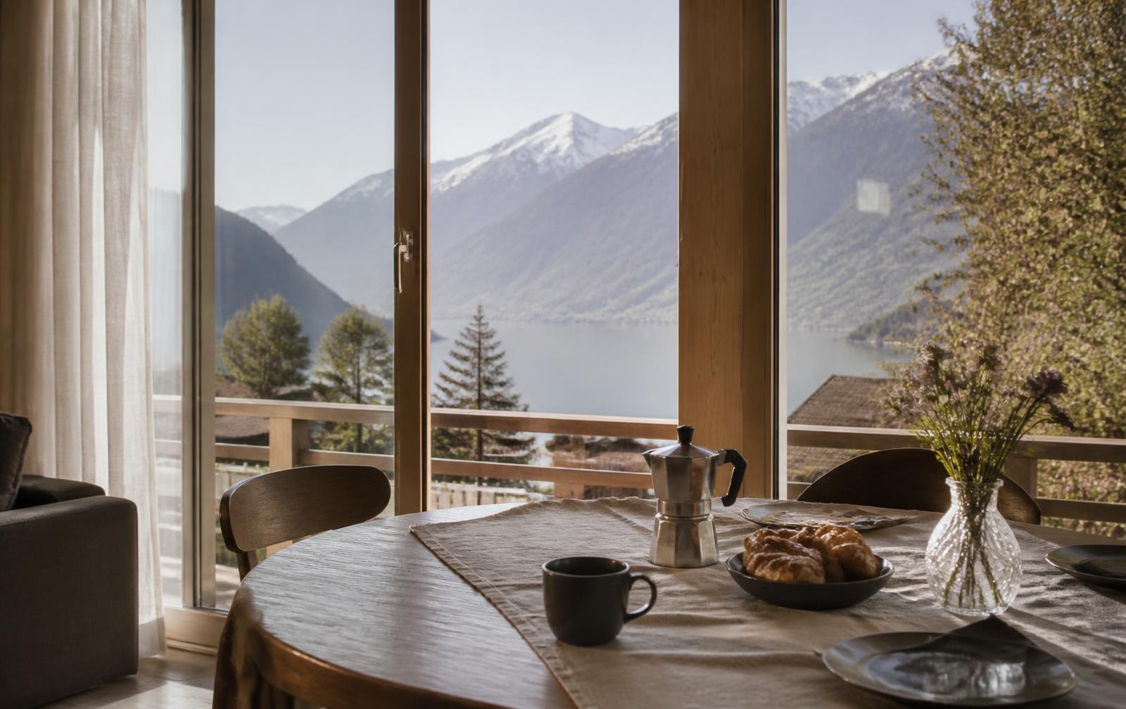 Gedeckter Holztisch in einer Ferienwohnung mit Blick durch grosse Fenster auf See und Berge