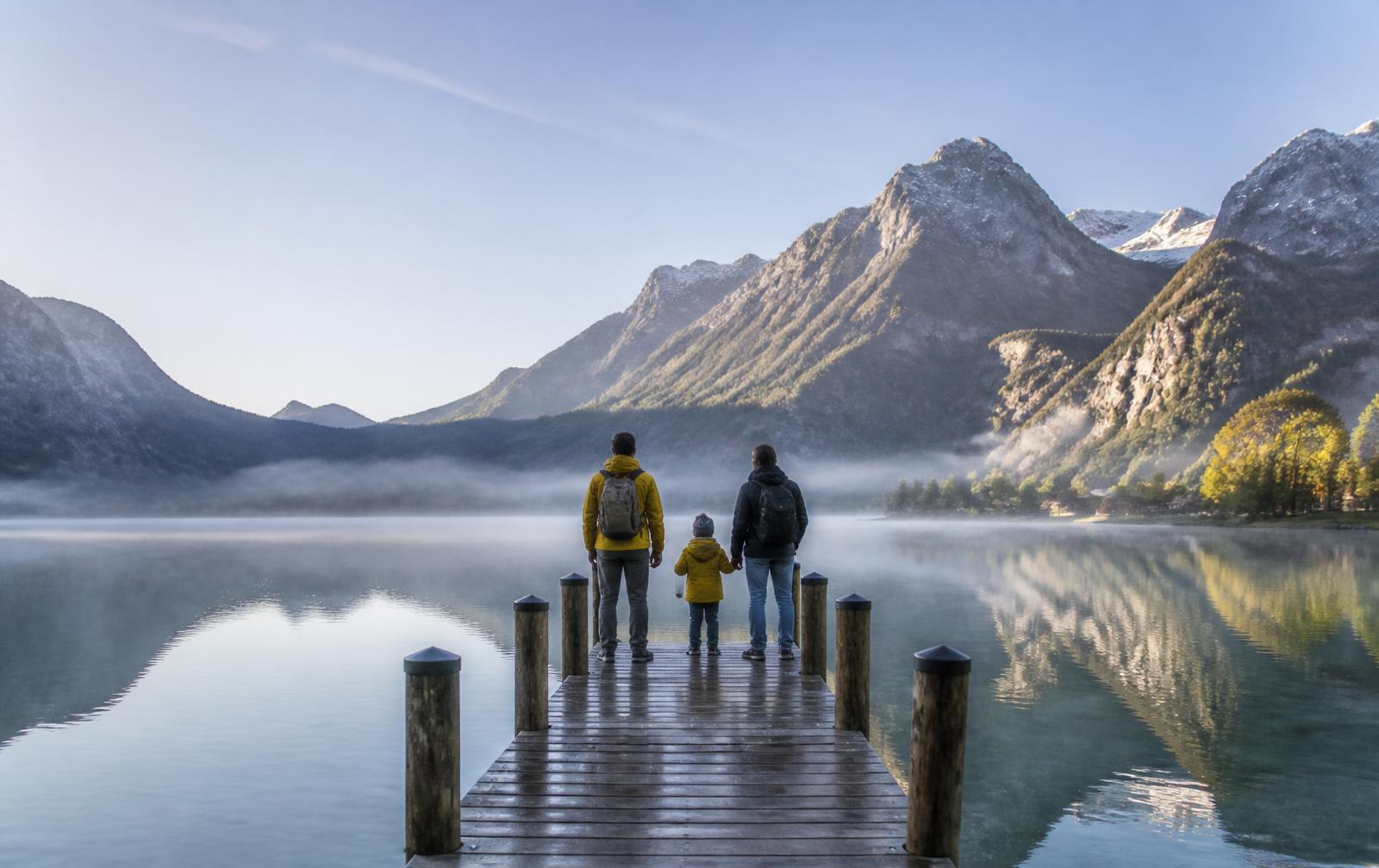 Familie steht am frühen Morgen auf einem Holzsteg an einem nebligen Bergsee